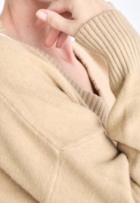 Beżowy sweter dzianinowy z prążkowanymi mankietami i dekoltem w kształcie litery V. Posiada miękką teksturę z delikatnym wzorem na tkaninie.