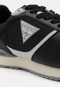 Zapatilla negra con un acento gris y tejido texturizado. Presenta un parche de logo plateado en forma de triángulo y cordones negros. Suela suave y ligera.