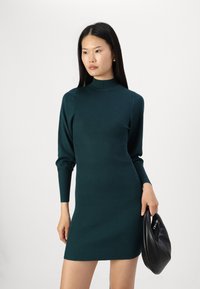 Rochie tricotată în teal închis, cu o textură canelată, guler înalt și mâneci lungi, asociată cu o poșetă texturată neagră.