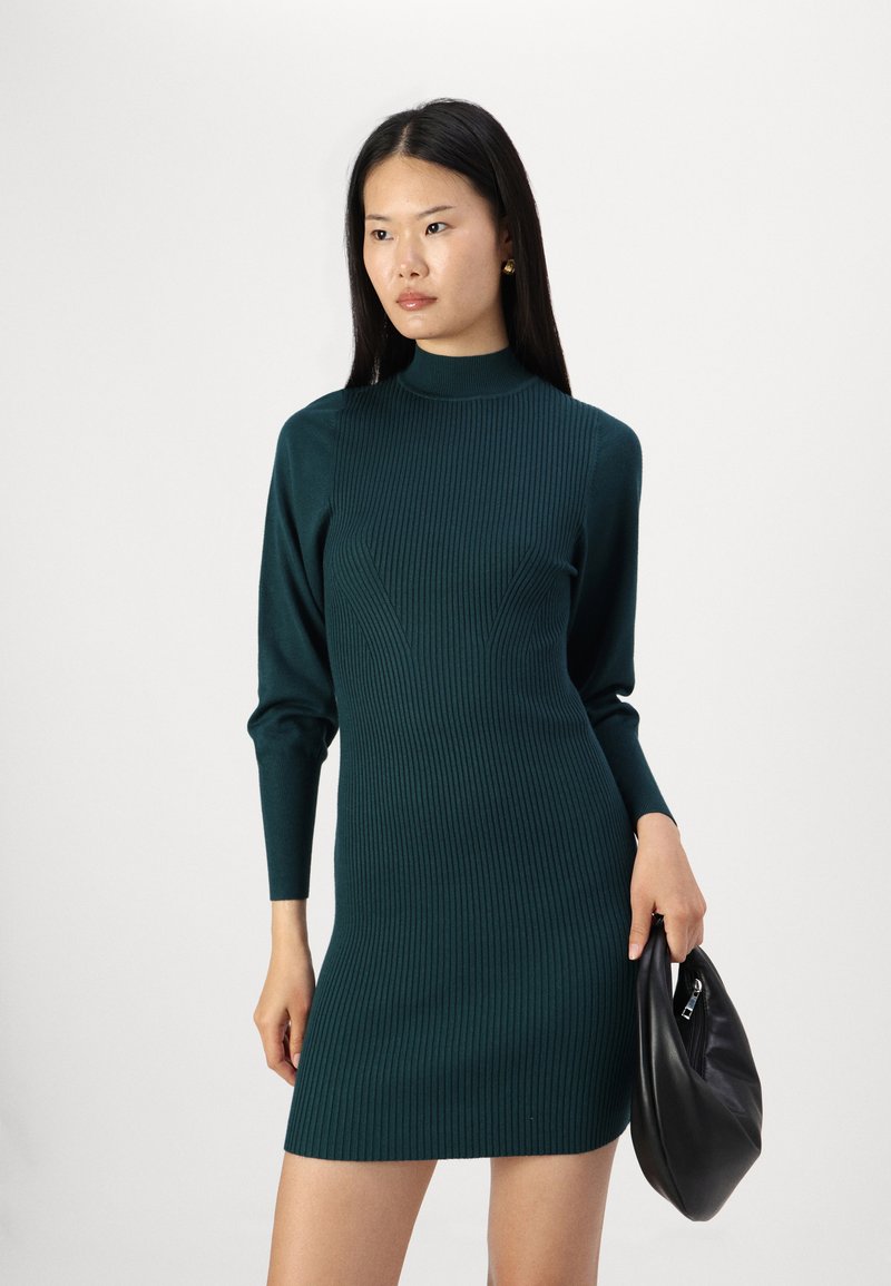 Gebreid jurkje in diep tealtint met een geribbelde textuur, hoge halslijn en lange mouwen, gecombineerd met een zwarte textuur clutch.