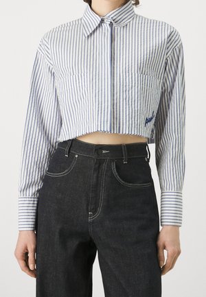 Vyötäröltä leikattu pitkähihainen paita, jossa on sinivalkoiset pystysuorat raidat ja kaksi rintataskua, yhdistettynä korkeavyötäröisiin tummansinisiin farkkuihin.