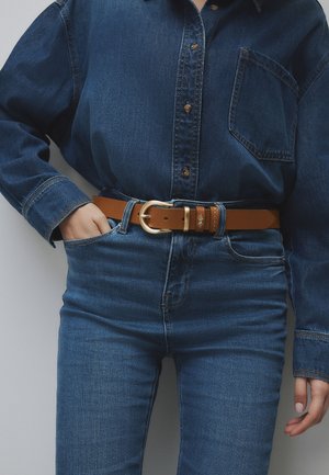 Persona che indossa una camicia di jeans blu scuro e jeans con cintura in pelle marrone con fibbia dorata, posando con una mano in tasca.