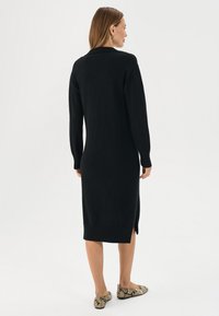 Abito nero in maglia con maniche lunghe, orlo a coste e spacchi laterali. Indossato con scarpe flat in stampa serpente, che mettono in evidenza una texture morbida.