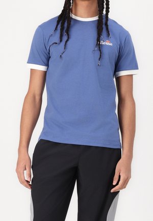 Personne portant un t-shirt bleu à manches courtes avec une bordure blanche et un pantalon noir, se tenant devant un fond blanc.