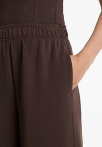 Rechte Hand in die Tasche von braunen, elastischen Bundhosen gesteckt, von der Taille bis zur Mitte des Oberschenkels vor einem weißen Hintergrund dargestellt.