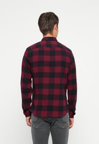 Hombre de espaldas vistiendo una camisa de cuadros roja y negra de manga larga y jeans grises, frente a un fondo blanco liso.