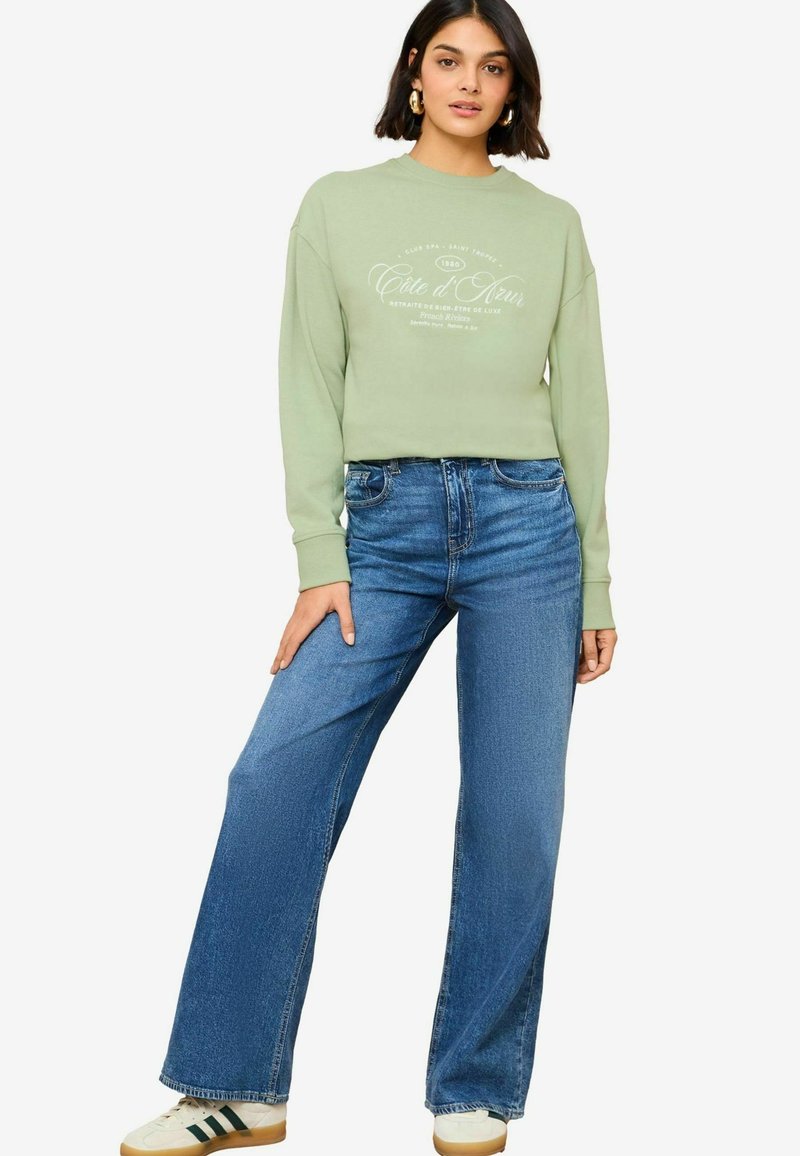Hele roheline lühike sweatshirt valge grafitextiga, koos kõrge vöökohaga siniste teksapükste ja valgete sneakersidega, millel on rohelised triibud.