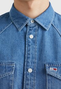 Jeansskjorta i ljusblått med spetskrage, framknappning, två bröstfickor och logotyp i röd, vit och blå på den högra fickan.