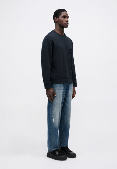 Άνδρας που στέκεται με μαύρη φούτερ, φθαρμένα μπλε τζιν και μαύρα αθλητικά παπούτσια μπροστά σε απλό λευκό φόντο.