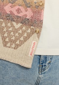 Mehrfarbiger, strukturierter Cardigan mit geometrischem Muster, beigem Grund, rosa und braunen Akzenten, ausgestattet mit drei Knöpfen und einem Etikett mit der Aufschrift "REVOLUTION."