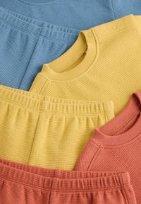 Ensembles doux en maille gaufrée composés de hauts et de pantalons à taille élastique, dans des couleurs bleu, jaune et rouille, disposés à plat pour présentation.