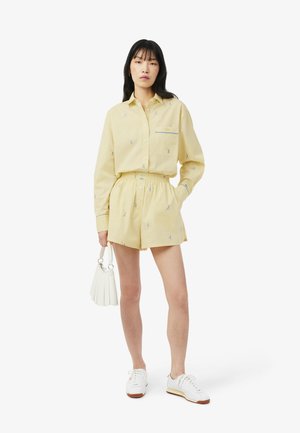 Femme portant une chemise boutonnée jaune pâle et un short à taille élastique assorti avec de petits motifs bleus, tenant un sac à main blanc et portant des baskets blanches.