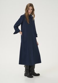 Rochie lungă, cu mâneci lungi, de culoare albastru închis, cu guler, închidere cu nasturi și buzunare laterale; combinată cu cizme negre până la genunchi. Textură de stofă netedă.