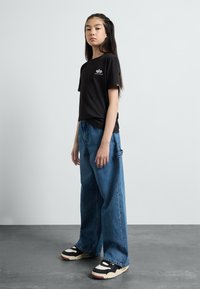 T-shirt en coton noir à manches courtes avec logo. Jean en denim bleu à patte large avec une poche latérale. Baskets noir et blanc avec des accents rouges.