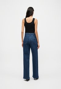 Γυναίκα με μακριά σκούρα μαλλιά, που φοράει μαύρο αμάνικο τοπ και ψηλόμεσα σκούρα μπλε παντελόνια με φαρδιά μπατζάκια, στέκεται γυρισμένη με την πλάτη σε λευκό φόντο.