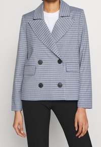 Dubbelsidig kavaj i blå och vit rutig mönster. Har stora svarta knappar och en bred krage. Skräddarsydd passform med sidofickor.