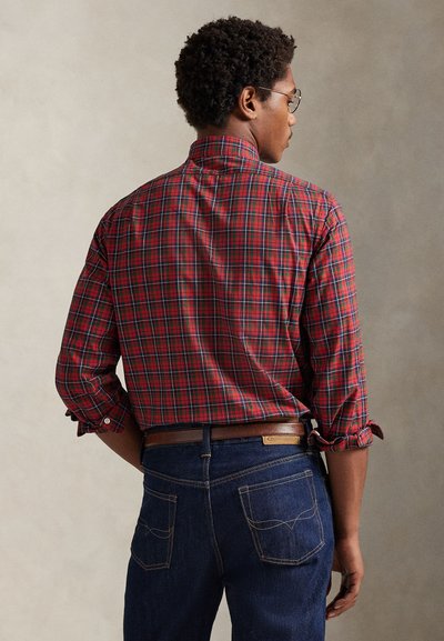 Czerwona i zielona flanelowa koszula w kratę z podwiniętymi rękawami, zestawiona z ciemnoniebieskimi dżinsami z tylnymi kieszeniami i brązowym paskiem.