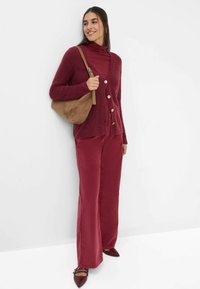 Burgunderfarbener Cardigan mit Knöpfen, über einem passenden Rollkragenpullover und weit geschnittenen Hosen. Braune Wildlederhandtasche und burgunderfarbene spitze Schuhe.