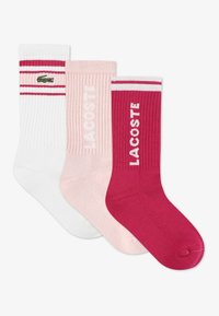 Τρία ζευγάρια ριμπ κάλτσες: λευκές με κόκκινες ρίγες, ροζ με λογότυπο "LACOSTE" και κόκκινες με λευκές λεπτομέρειες. Διαθέτουν απαλό βαμβακερό υλικό.