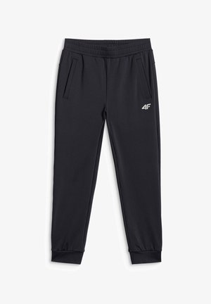Lachend kind staand, draagt een rood shirt met korte mouwen, zwarte sportbroek en zwarte sneakers tegen een effen grijze achtergrond.
