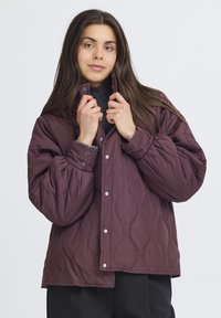 Burgundy quiltad jacka med hög krage, tryckknappsstängning och långa ärmar. Har ett texturerat mönster och en lös passform.