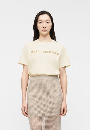 Kvinde med langt mørkt hår iført en beige Karl Lagerfeld T-shirt og et højtaljet beige mesh-nederdel, stående mod en ensfarvet hvid baggrund.