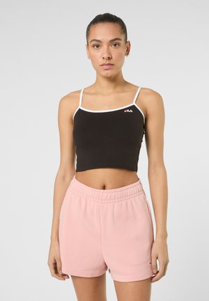 Frau mit zurückgebundenen Haaren, die ein schwarzes, kurzes Tanktop mit weißem Rand und hellrosa Shorts mit elastischem Bund trägt, steht vor einem einfarbigen Hintergrund.