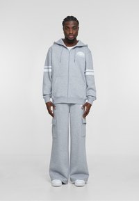 Grå zip-up hoodie med et logo, der har hvide striber på ærmerne, parret med brede cargo bukser med lommer og hvide sneakers.