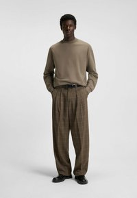 Pantaloni a scacchi marroni con pinces, abbinati a un top beige a maniche lunghe con scollo rotondo e branding sottile. Cintura e scarpe nere.