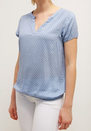 Femme portant un chemisier bleu clair à manches courtes avec de petits motifs blancs et un pantalon blanc, debout devant un fond uni.