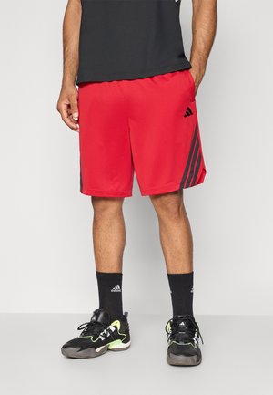 Mand iført røde Adidas shorts, sorte Adidas sokker og sorte sneakers med neon grønne accenter, står mod en ensfarvet baggrund.