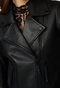 Chaqueta de motociclista de cuero negro que presenta un cuello con muescas, cierre frontal con cremallera y adornos. Tiene dos bolsillos con cremallera y una capa inferior de encaje texturizado.