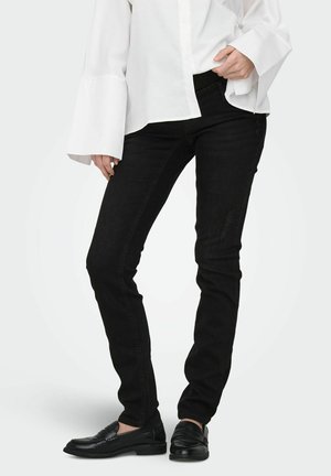 Osoba nosiaca čierne úzke džínsy, čierne mokasíny a bielu blúzku s dlhými rukávmi a širokými manžetami, stojaca pred jednoduchým pozadím.