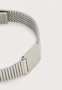 Ασημένιο μεταλλικό βραχιόλι ρολογιού με πλεκτό σχέδιο, με ένα λείο κούμπωμα χαραγμένο με τη λέξη "HUGO". Η υφή είναι κομψή και γυαλιστερή.