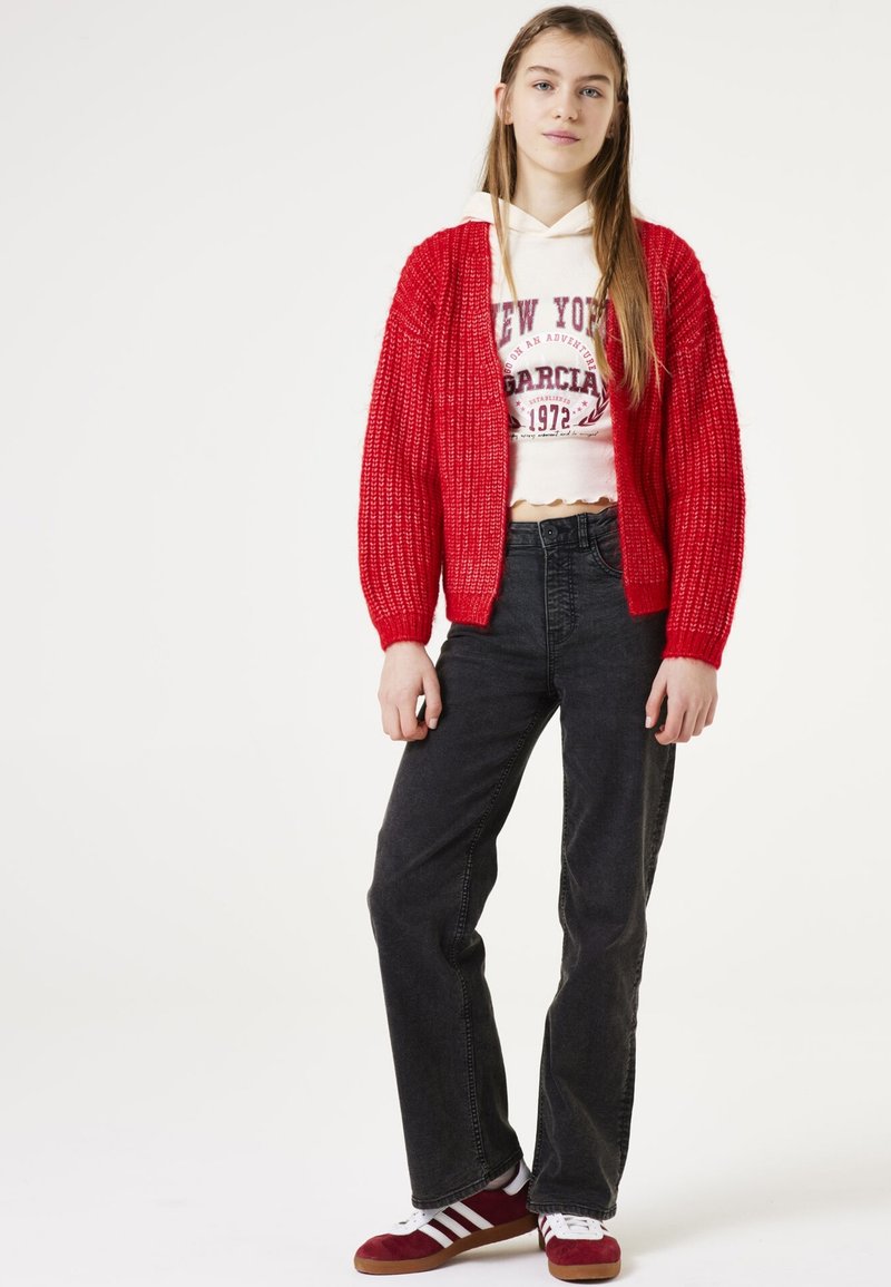 Rode gebreide cardigan over een witte cropped hoodie met print, gecombineerd met zwarte denim jeans en rode sneakers met witte strepen.