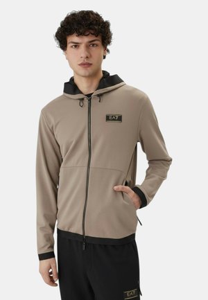 Giovane uomo che indossa una felpa beige con zip e logo EA7 e pantaloni neri con logo abbinato, in piedi con una mano in tasca.