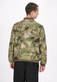 Kamouflagemönstrad jacka i grönt, brunt och beige. Har en krage och ribbade ärmslut. Tyget har en slät yta och en avslappnad passform.