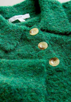 Groene geztructureerde wollen kledingstuk met drie grote gele knopen en een kraag. Etiket zichtbaar aan de binnenkant, dat het merk "NEXT" aangeeft.