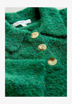 Groene geztructureerde wollen kledingstuk met drie grote gele knopen en een kraag. Etiket zichtbaar aan de binnenkant, dat het merk "NEXT" aangeeft.