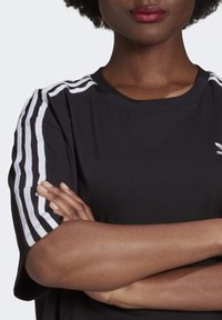 Black short-sleeve t-shirt with white three-stripe design on shoulders and small logo. Material appears soft, with a classic crew neckline.