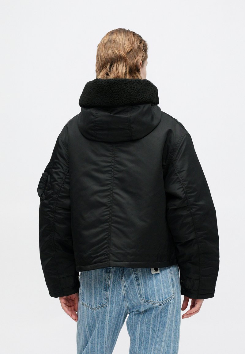 Schwarze gepolsterte Jacke mit Kapuze, die einen strukturierten Kunstpelzkragen hat. Das Design umfasst weite Ärmel und eine verkürzte Länge, getragen über hellblauen gestreiften Jeans.