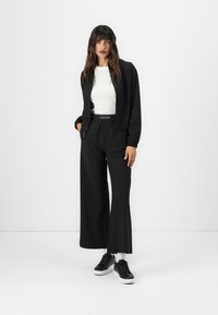 Zwarte zip-up jas over een wit shirt, gecombineerd met losse zwarte broeken met een Calvin Klein tailleband en zwarte sneakers.