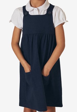 Marineblauwe katoenen jurk met een geplooide rok, voorzien van twee zakken aan de voorkant en knopen op de schouders. Witte blouse met kraag eronder.