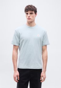 Νέ ος άνδρας που φοράει κοντομάνικο μπλουζάκι ανοιχτού μπλε με υφή και μικρό λογότυπο στο στήθος, συνδυασμένο με μαύρο παντελόνι, κοιτάζοντας μπροστά.