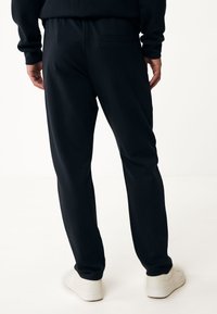 Pantalones de chándal negros con un corte holgado, cinturilla elástica y un único bolsillo trasero, combinados con zapatillas blancas sobre un fondo claro.
