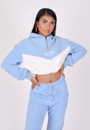 Jeune femme aux longs cheveux foncés portant un sweat à capuche court bleu et blanc assorti avec un pantalon de survêtement bleu, debout, les mains sur les hanches.