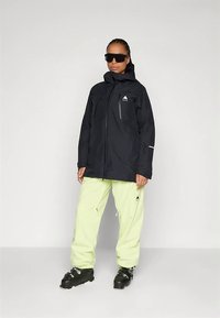 Person in einer schwarzen Winterjacke, blassgelben Schneehosen, schwarzen Skischuhen und großen schwarzen Sonnenbrillen, steht vor einem schlichten Hintergrund.