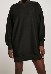 Abito nero a coste oversize con maniche lunghe, scollo a V e polsini. Tessuto morbido, che raggiunge la metà della coscia, progettato per comfort e facilità.