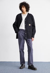 Black button-up shirt with a chest pocket, paired with light gray pants. Model wears black shoes and a silver chain necklace.