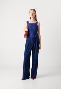 Haut sans manches bleu marine avec un motif texturé et des embellissements, assorti à un pantalon ample bleu marine et des escarpins à bout pointu rouges, tenant un sac rouge.